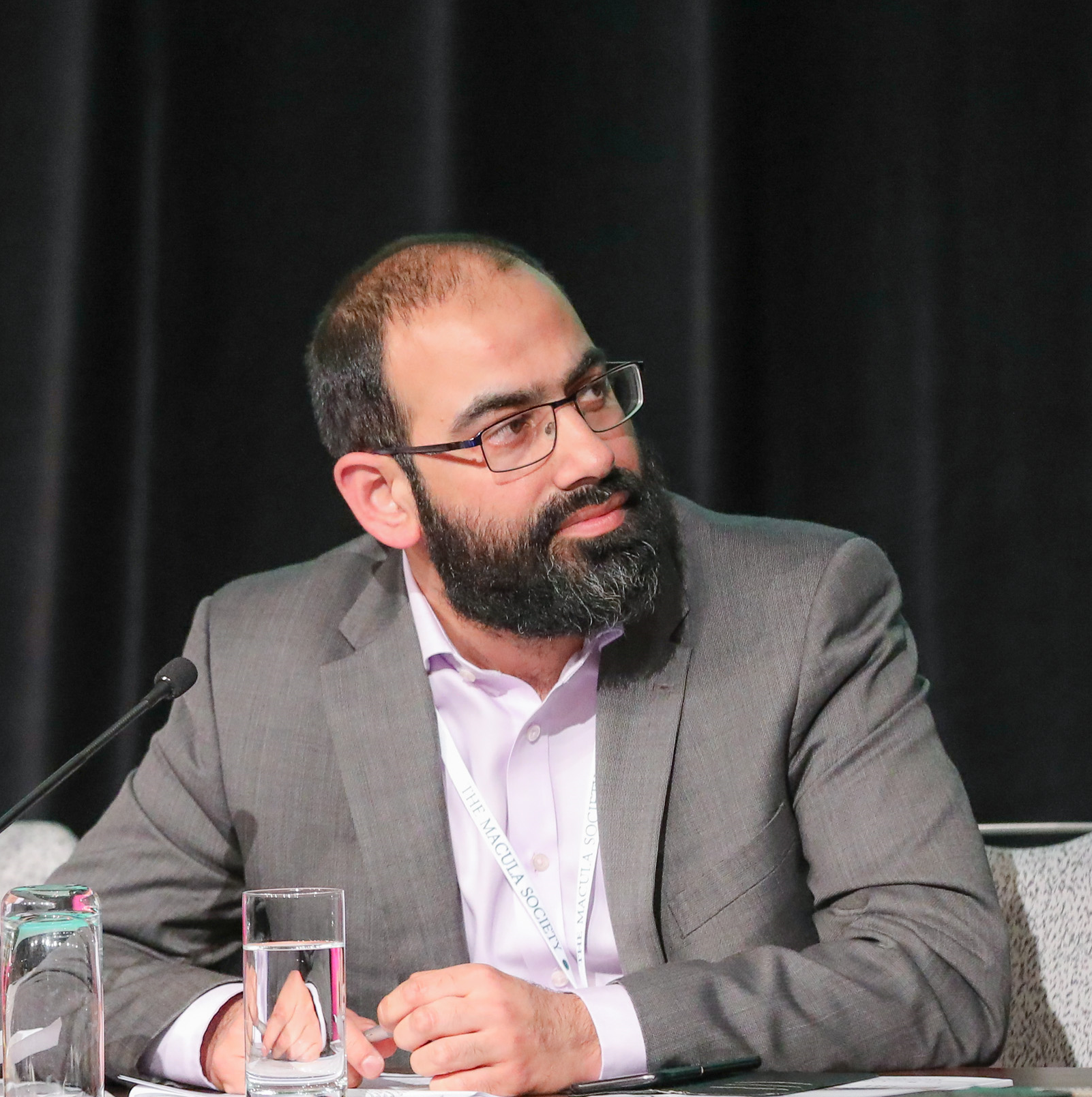 Omar Mahroo. He has dark hair and a beard. He is wearing glasses and a grey suit. 